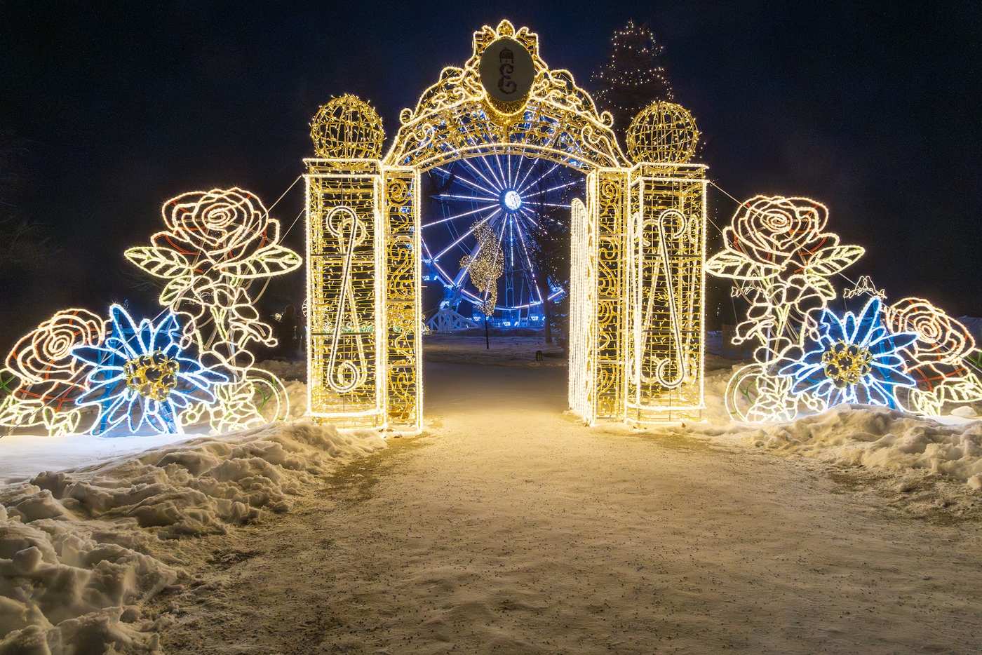 An ornate light gate decorated with roses and blue flowers, a lit ferris wheel behind it