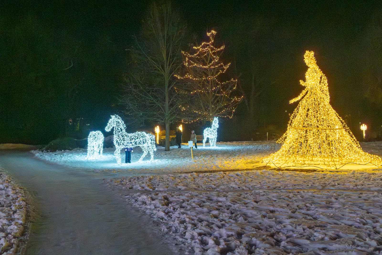 A tall crinoline dress rendered in golden light beside white horse sculptures on a snow-covered lawn