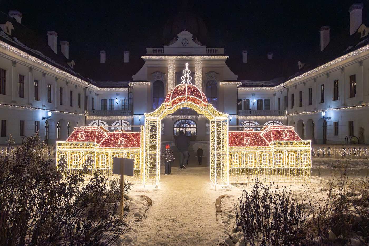 The illuminated baroque facade of the Royal Palace of Gödöllő behind a gate of light shaped like stacked gift boxes, snow on the ground